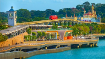 ภาพยามกลางวันของรถไฟฟ้าสีแดงที่ตัดผ่านเส้นทางเดิน Sentosa Boardwalk โดยมีสวนสนุก Universal Studios Singapore เป็นฉากหลัง