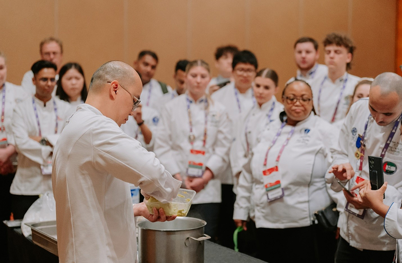 Senior chef demonstrates dish preparation to younger chefs during WCC 2024.