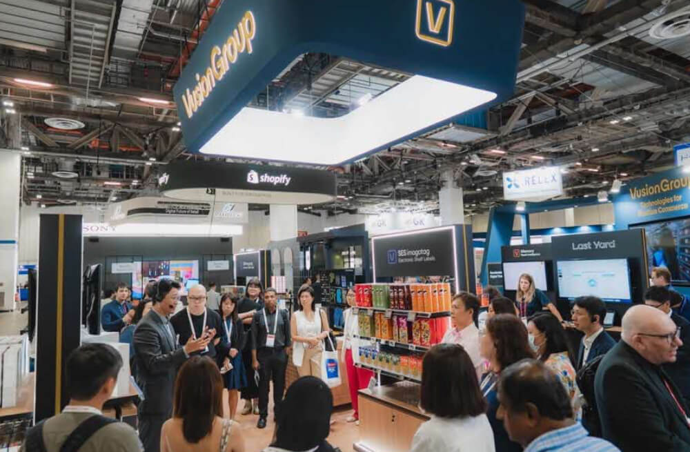 A group at Retails Big Show listens to a speaker near product displays and company booths.