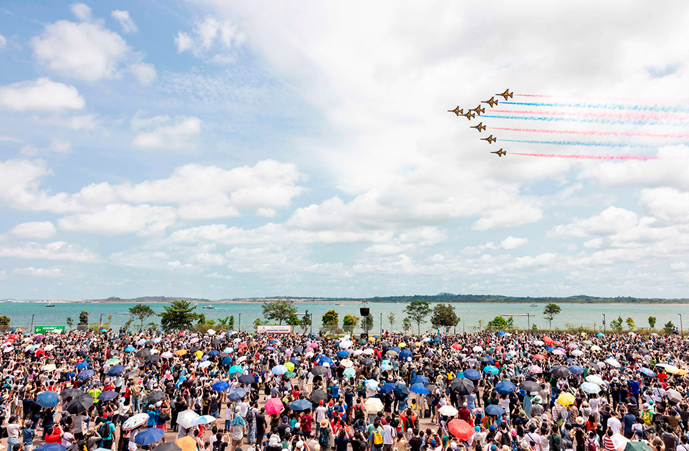 Colorful smoke behind 8 fighter planes during aerial stunt at Singapore Airshow 2024, crowd watching.