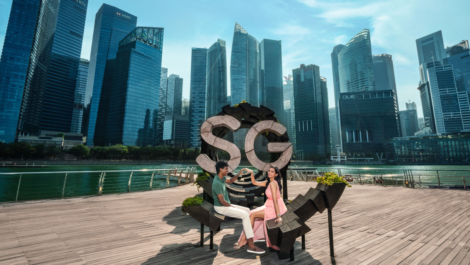 View of a couple posing next to Here is SG sculpture with Singapore’s Marina Bay skyline