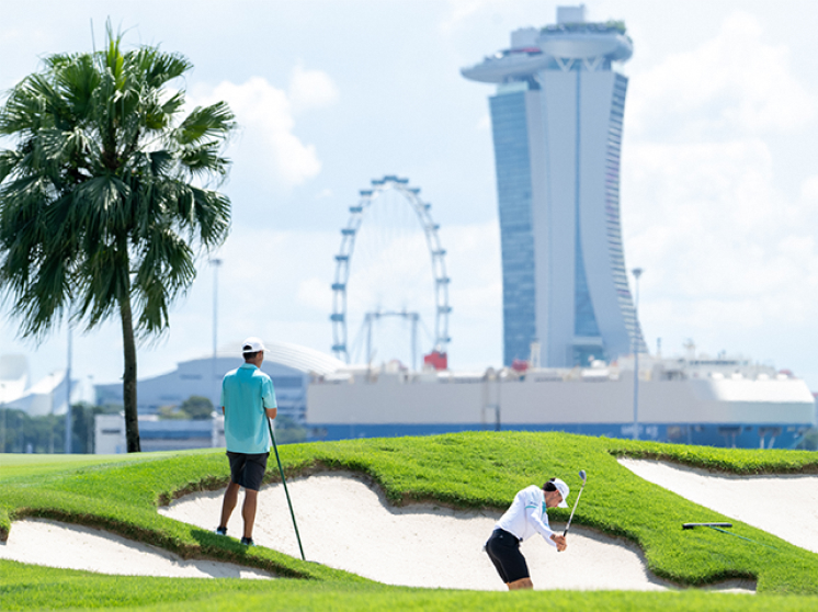 Golfers on a green with sand traps, featuring the Singapore Flyer and Marina Bay Sands skyline.