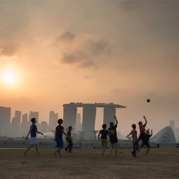 Jungs beim Fußballspielen auf der Marina Barrage in der Dämmerung, mit der verschwommenen Silhouette der Marina Bay im Hintergrund