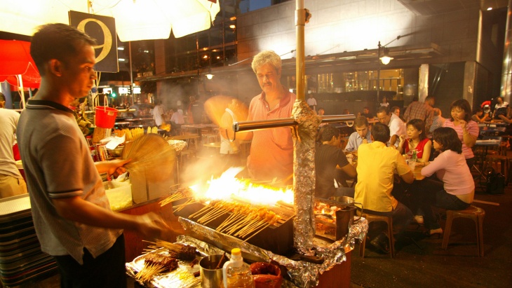 A tourist looking at a street hawker making satay over a flame at Lau Pa Sat