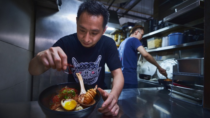 Portrait shot of an owner from A Noodle Story garnishing a bowl of wanton noodles 