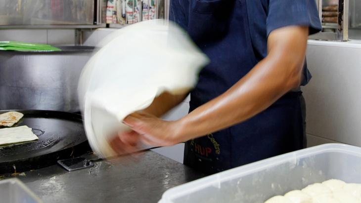 Close up of a man pulling dough to make <i>roti prata</i>.