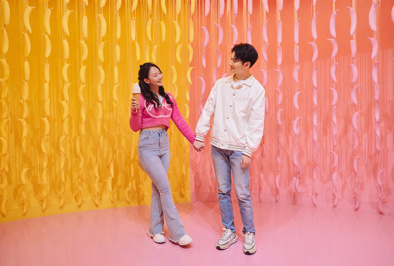 A smiling couple holding hands and enjoying ice cream inside the vibrant yellow and pink Banana Maze at the Museum of Ice Cream.