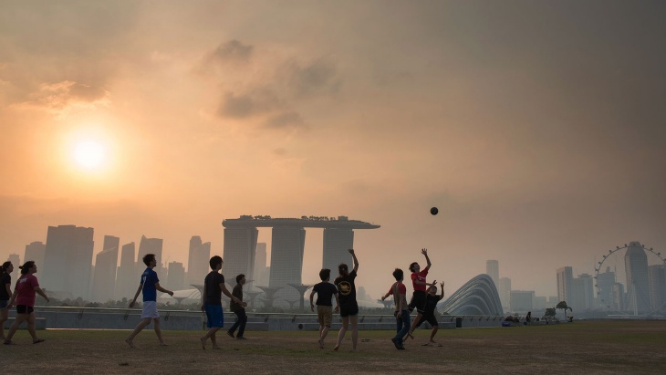 Sekumpulan anak lelaki bermain sepak bola di Marina Barrage saat senja, dengan siluet Marina Bay yang berkabut di belakangnya