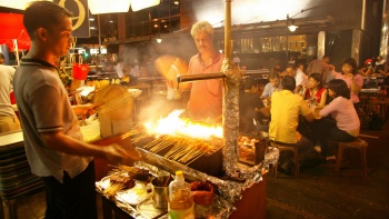 Foto malam eksterior para pria membakar sate dengan pengunjung yang sedang menikmati santapan kuliner