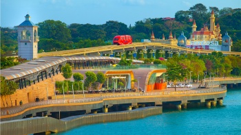 ภาพยามกลางวันของรถไฟฟ้าสีแดงที่ตัดผ่านเส้นทางเดิน Sentosa Boardwalk โดยมีสวนสนุก Universal Studios Singapore เป็นฉากหลัง