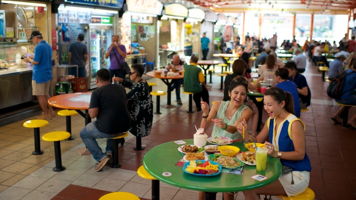 ผู้คนนั่งรับประทานอาหารกันที่ Maxwell Food Centre โดยมีแผงขายอาหารอยู่ฝั่งเดียวกัน