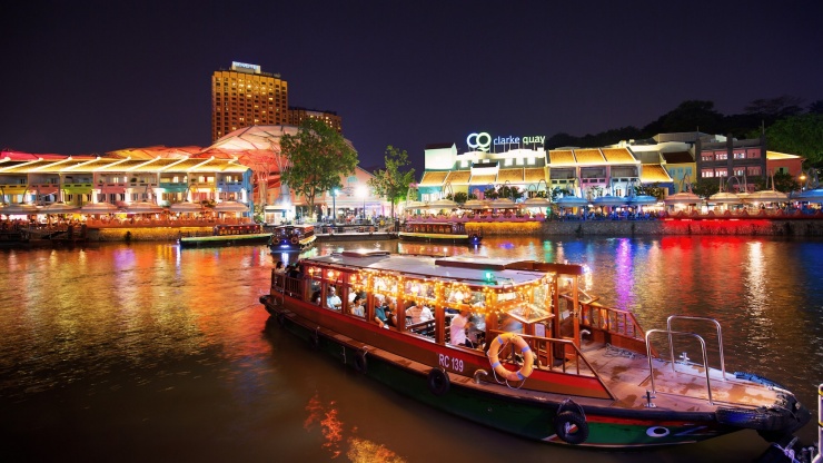 เรือบัมโบ๊ทที่จอดเลียบแม่น้ำสิงคโปร์ในยามค่ำคืน และมีป้าย Clarke Quay ปรากฏเป็นฉากหลัง