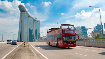 รถบัสสีแดงเปิดหลังคาที่ให้บริการ DuckTours กำลังแล่นอยู่บนถนนโดยมี Marina Bay Sands เป็นฉากหลัง