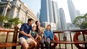 ครอบครัวสนุกสนานบนเรือนำเที่ยว Singapore River Cruise โดยมีย่านธุรกิจใจกลางเมืองเป็นฉากหลัง